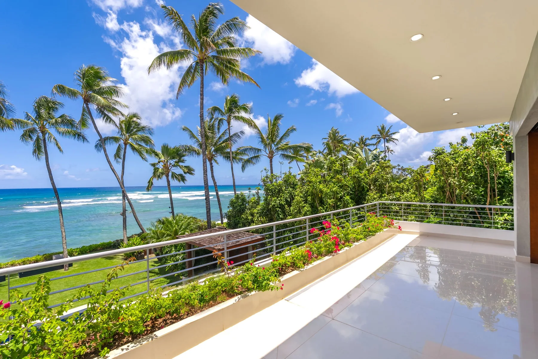 A covered balcony overlooks palm trees, greenery, and the ocean.
