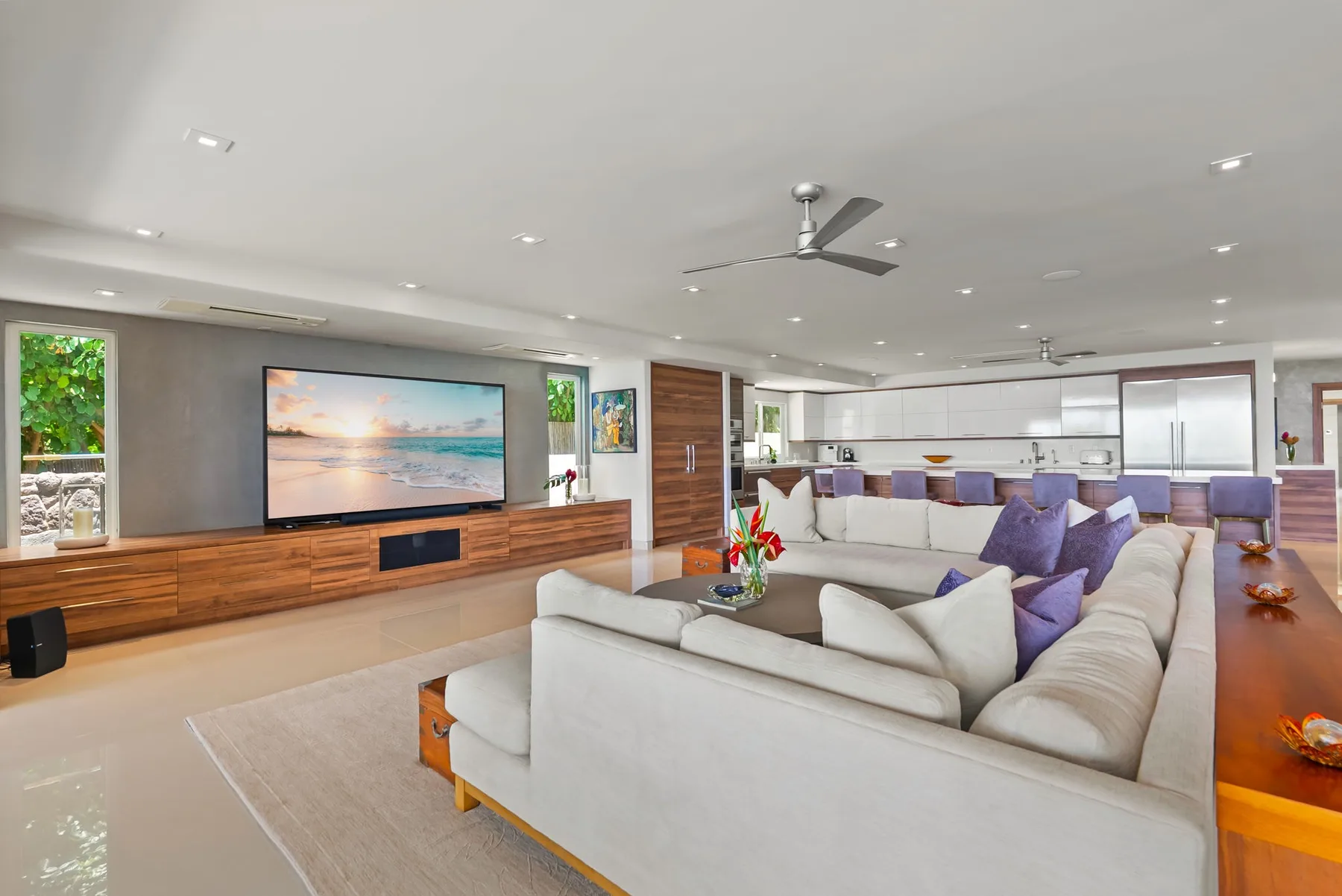 A spacious living room with light-colored seating, a television, and an open kitchen.