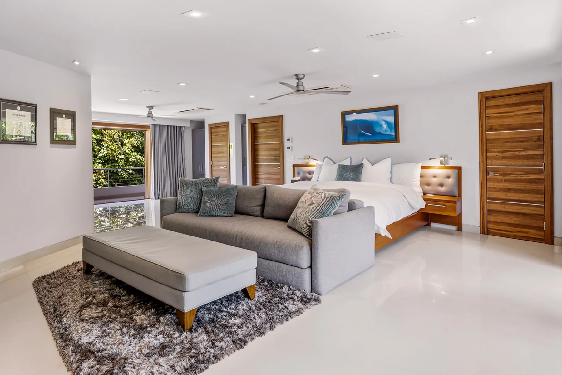 A bright bedroom with a large bed, gray seating, and a window overlooking greenery.