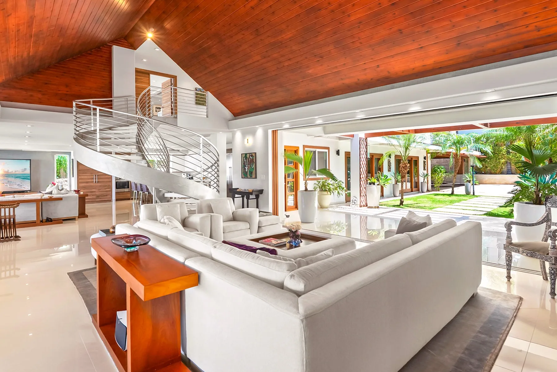 A bright living room with white seating and a curved staircase beneath a wood ceiling.