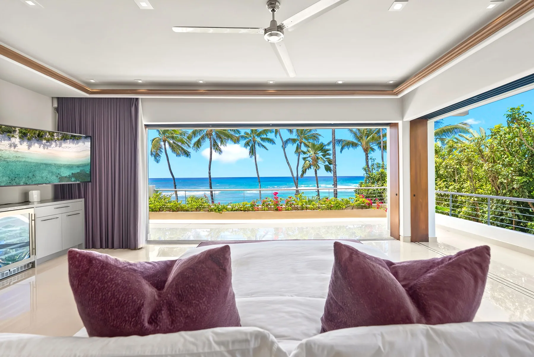 A bright bedroom opens onto a wide ocean view framed by palm trees.