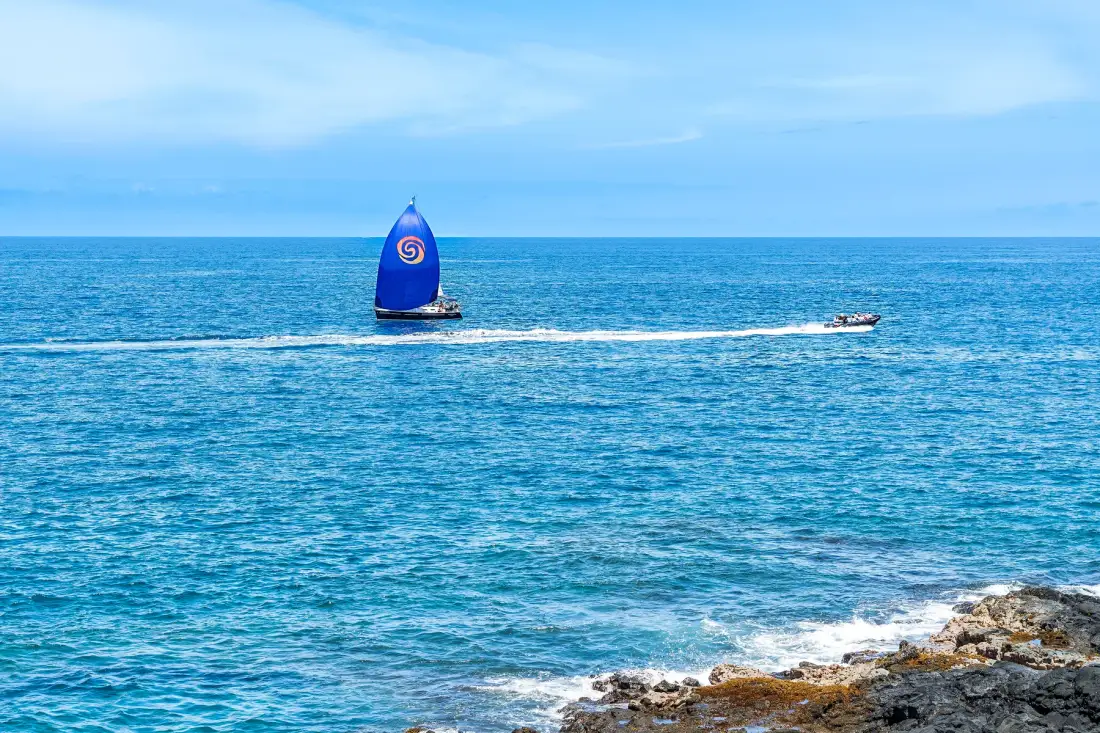Un velero azul navega por el mar cerca de una costa rocosa.