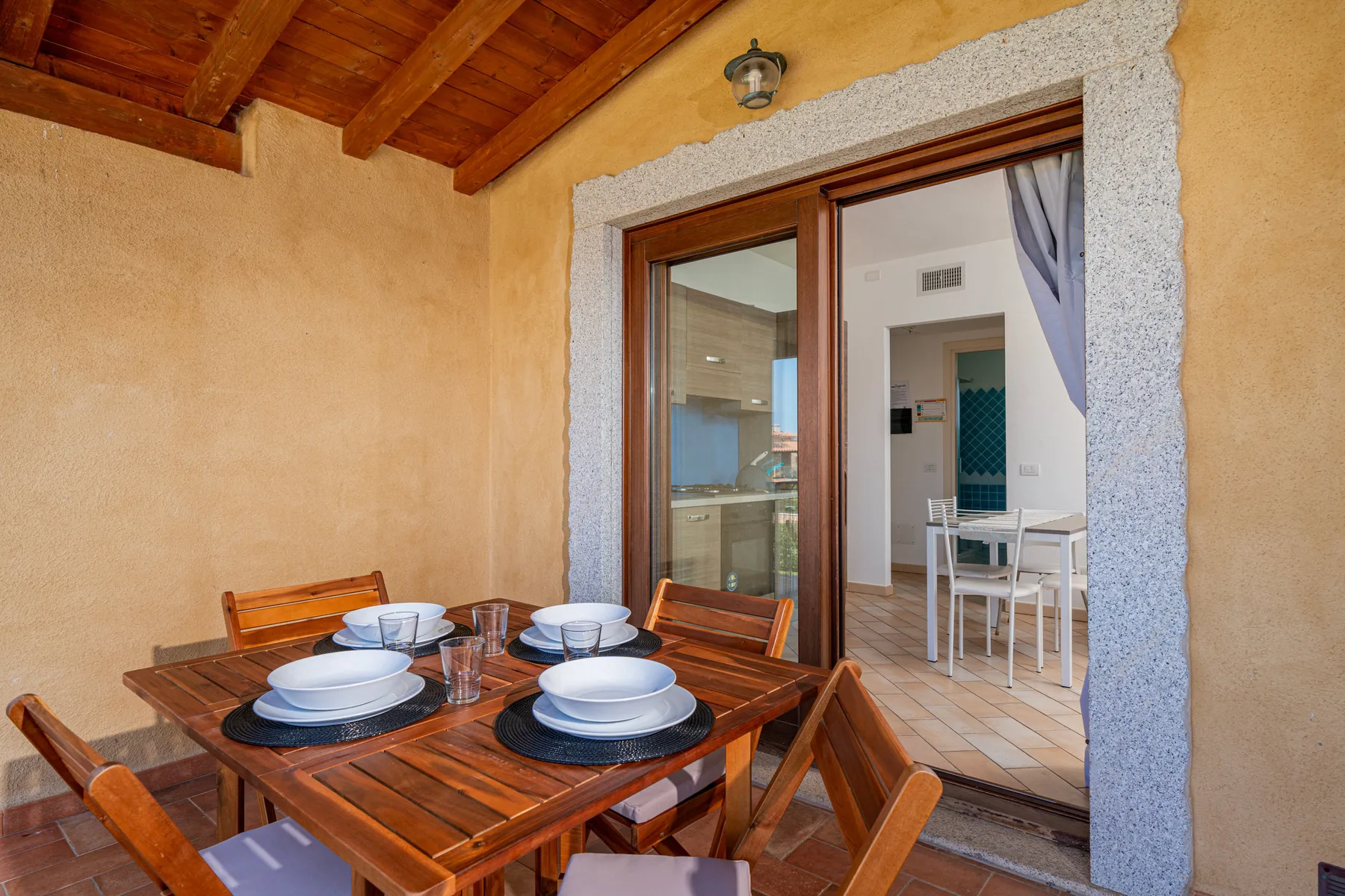 Una mesa de comedor de madera y asientos están en un patio cubierto junto a puertas de vidrio abiertas.