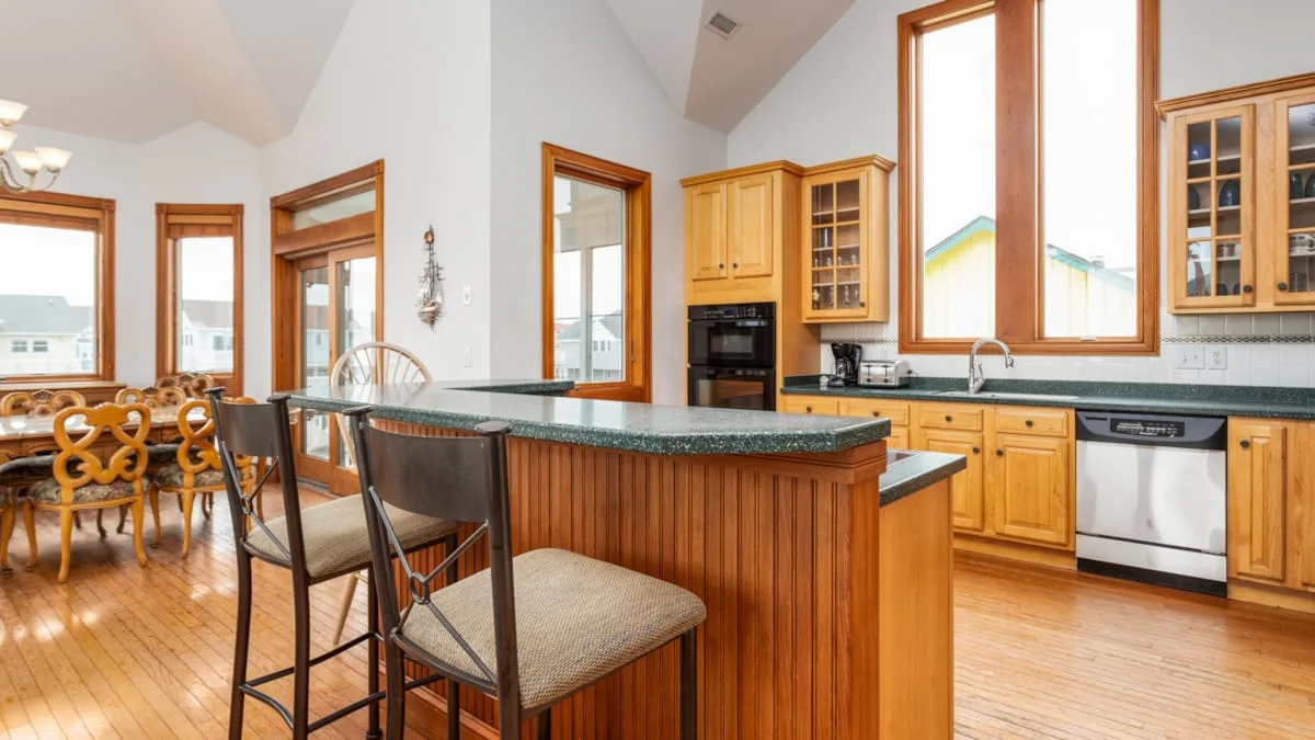 Una cocina luminosa tiene una isla de madera, asientos altos y ventanas grandes.