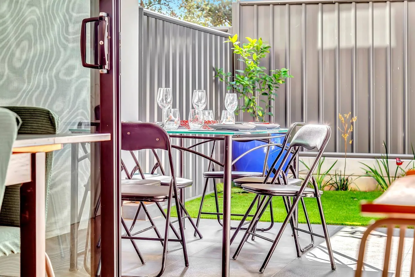 A patio seating area has a table set with glasses beside a grassy yard.