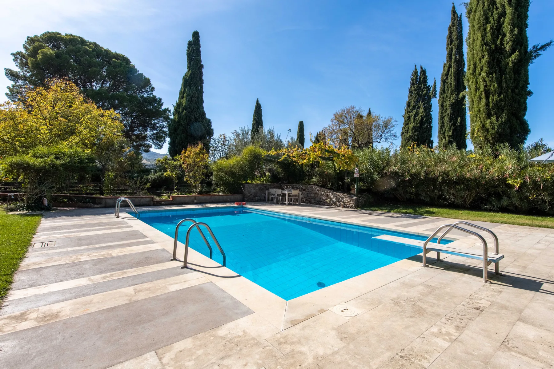 Una piscina al aire libre está junto a una terraza pavimentada y vegetación.