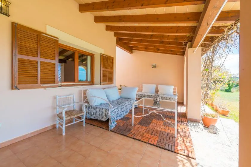 Un patio cubierto con asientos, una mesa pequeña y ventanas con contraventanas de madera.