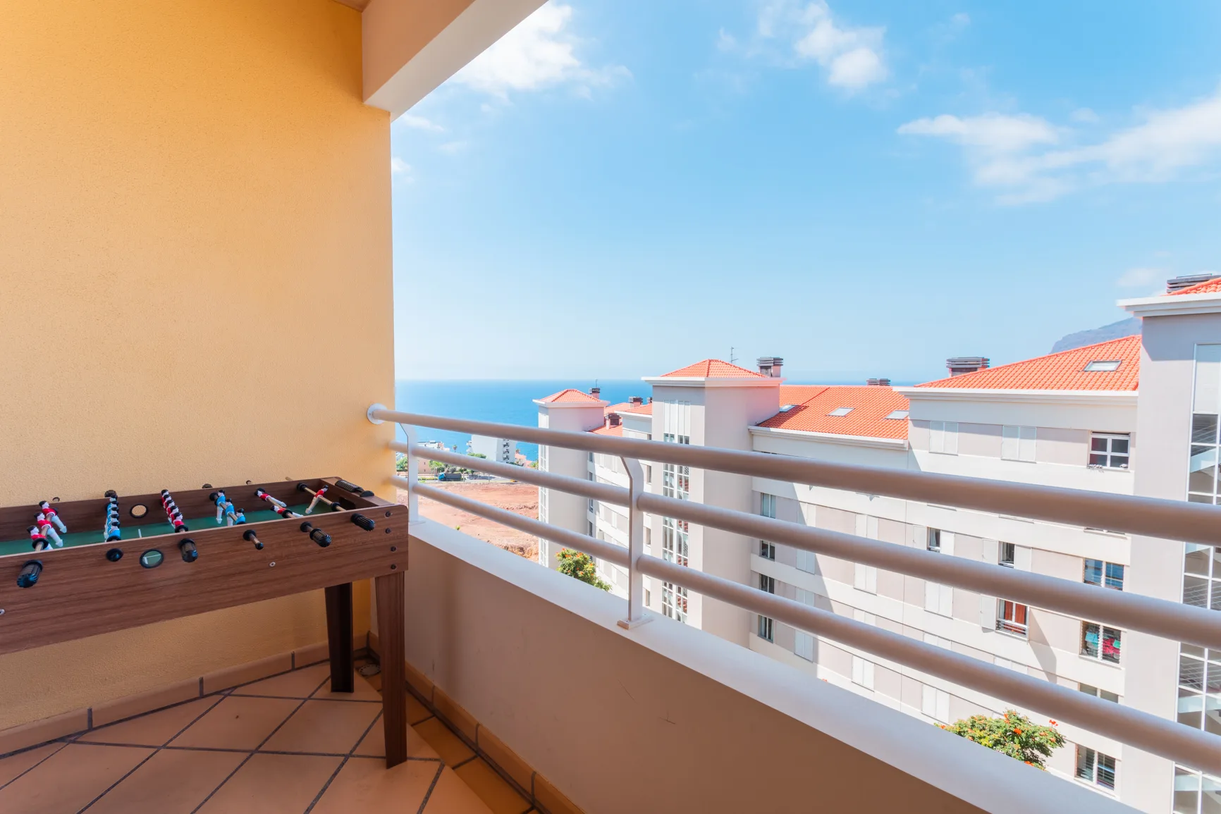 A balcony overlooks nearby buildings and the ocean, with a foosball table in the foreground.