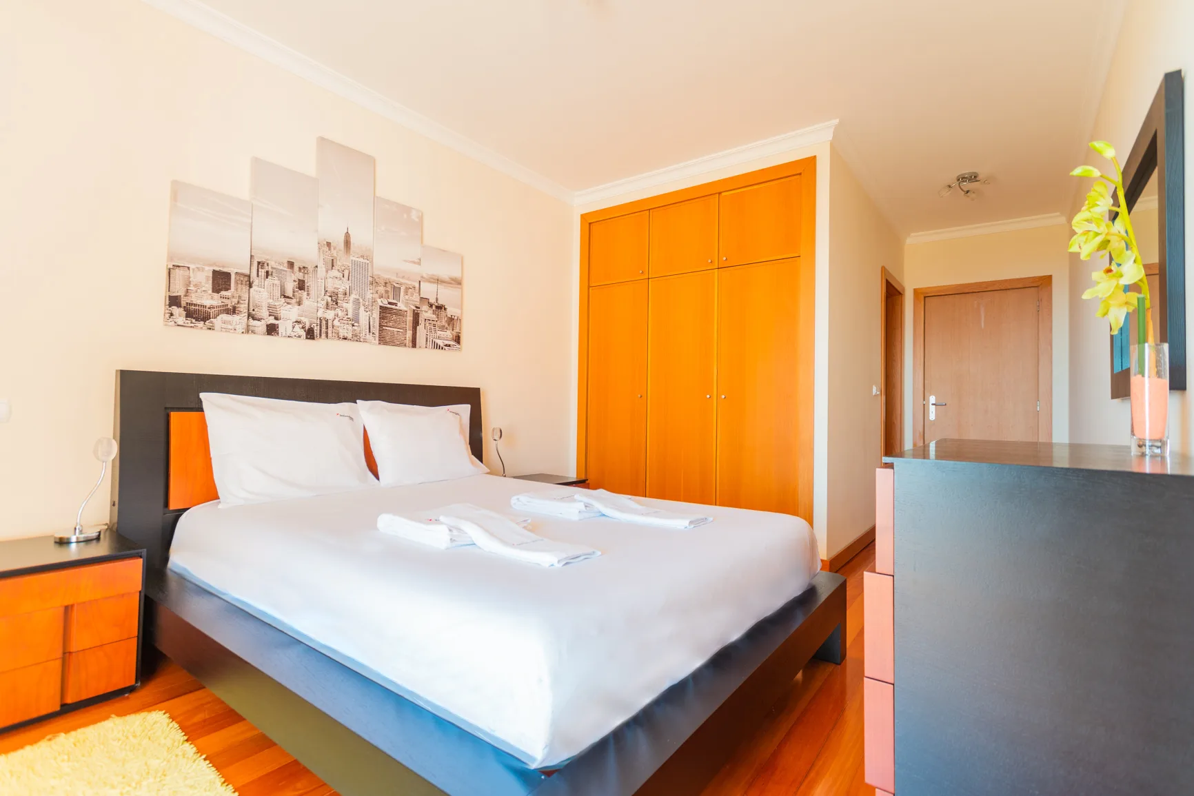 A bedroom with a double bed, orange wardrobe, and framed cityscape artwork.