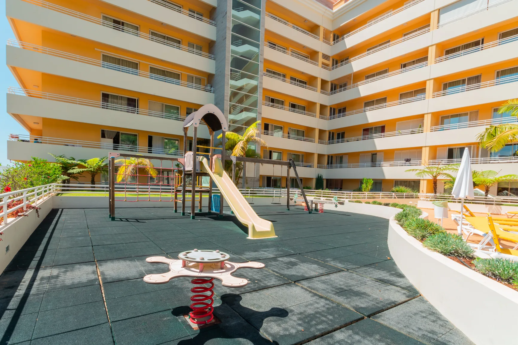 An outdoor playground with a slide and spring rider sits beside a multistory building.