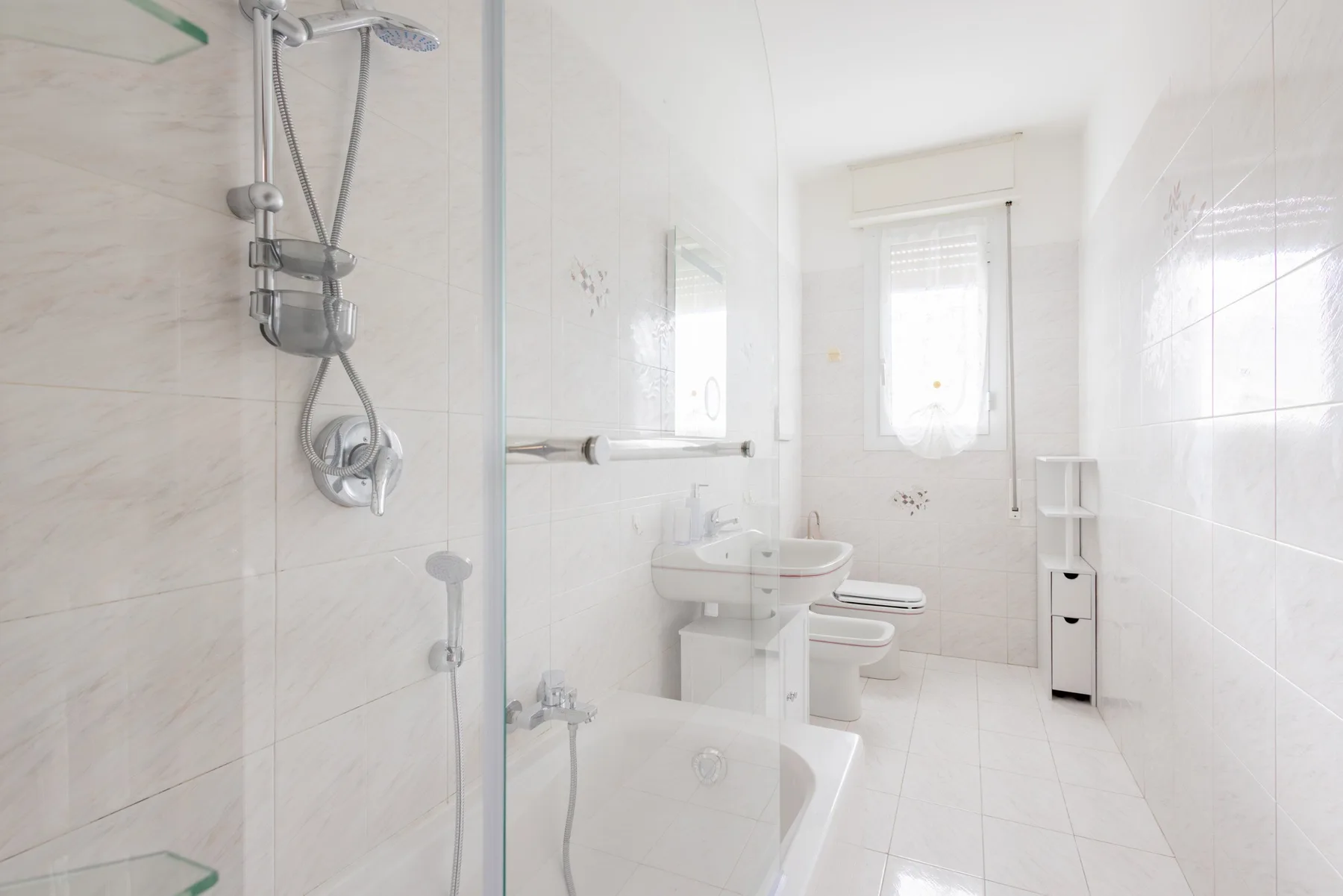 A bright tiled bathroom with a shower, sink, and window.