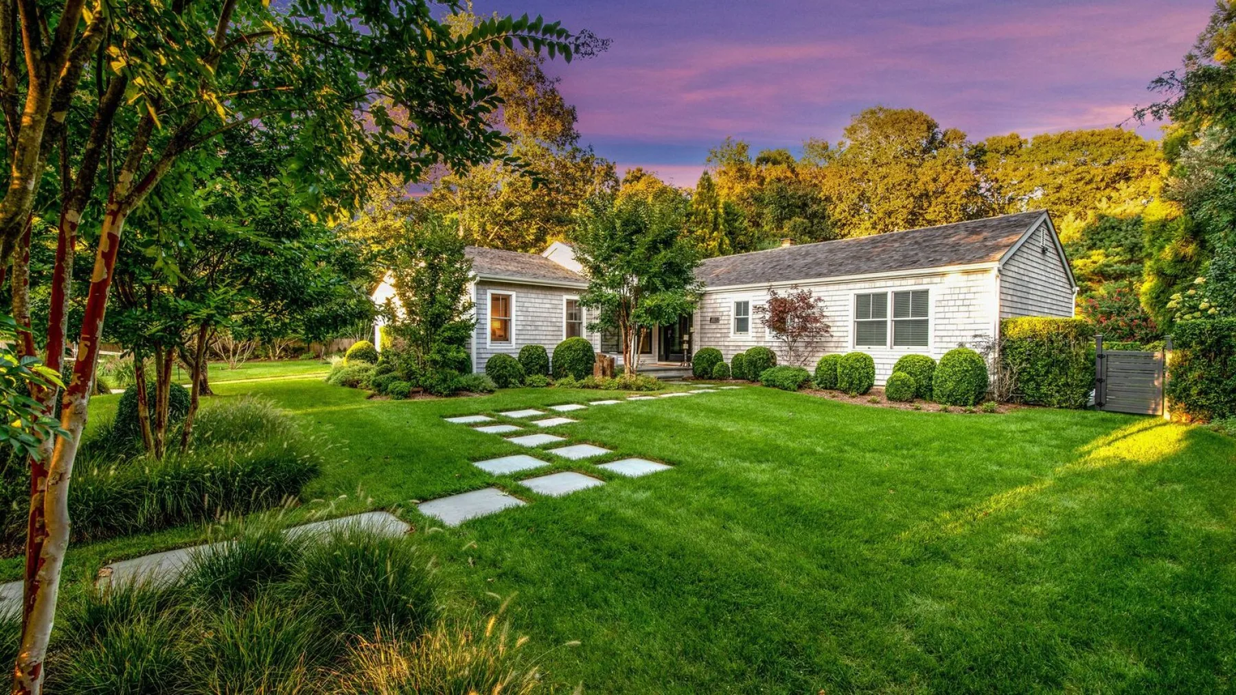 Una casa blanca junto a un césped verde, con piedras de paso y árboles.