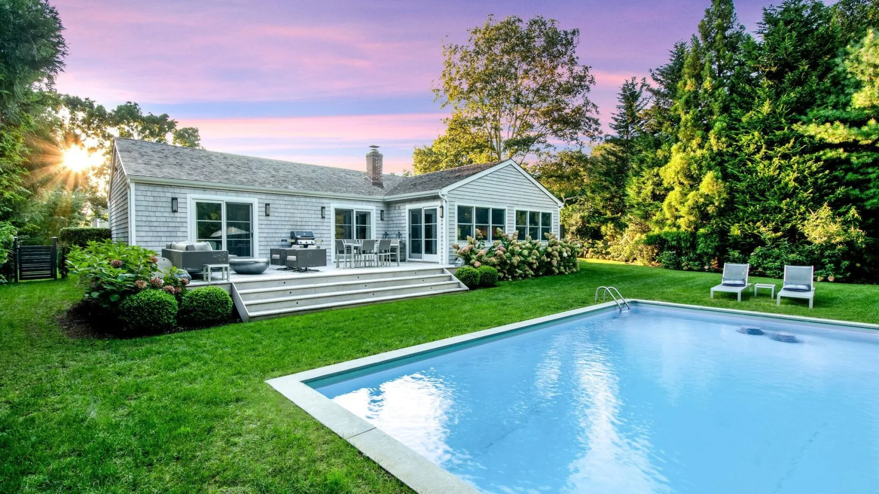 Una casa y una piscina están junto al césped, con árboles al fondo.