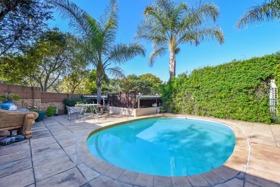 An outdoor swimming pool sits in a paved patio surrounded by greenery.