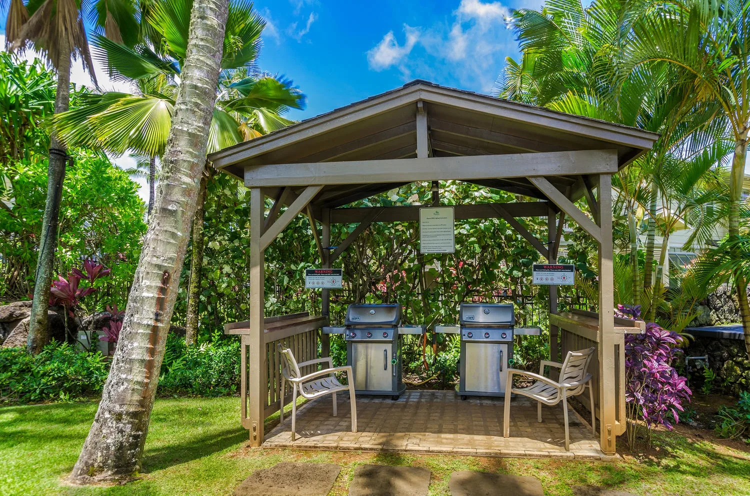 An outdoor cooking area under a wooden shelter, surrounded by tropical plants.