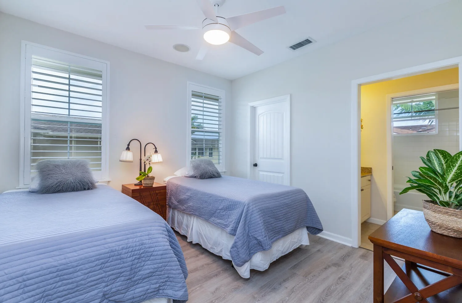 A bright bedroom with two beds, shuttered windows, and a ceiling fan.