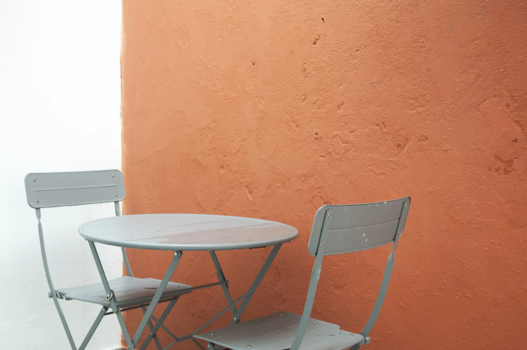 A small outdoor seating area with a round table and metal chairs beside an orange wall.