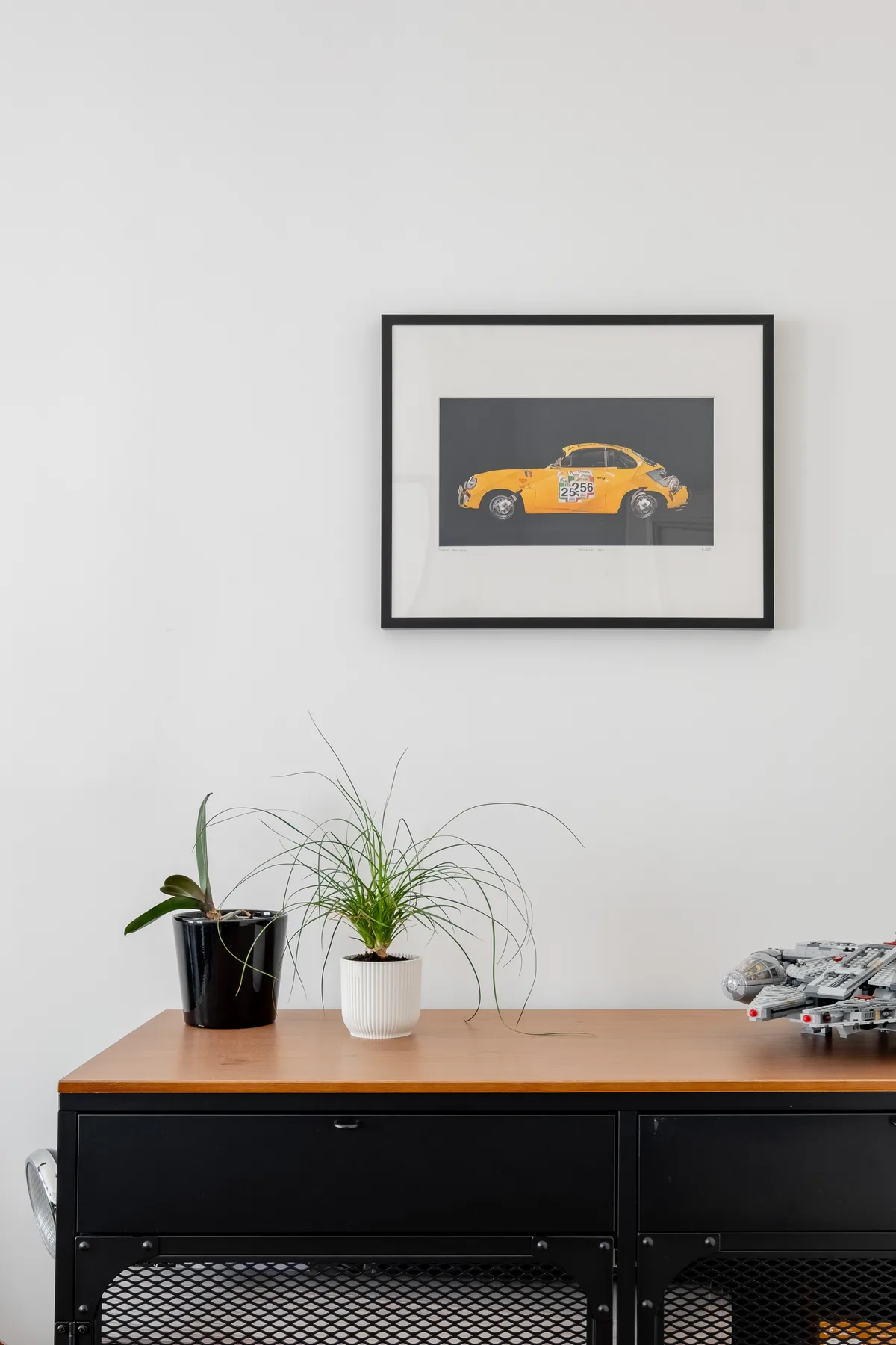 Un mueble negro con plantas en macetas y un cuadro de un auto sobre una pared blanca.