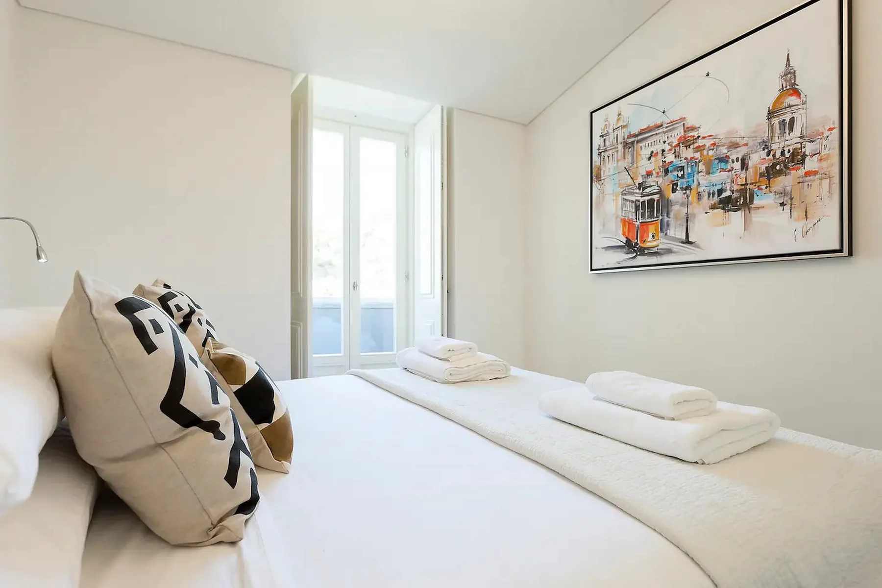 Un dormitorio luminoso con ropa de cama blanca, toallas dobladas y una obra enmarcada.
