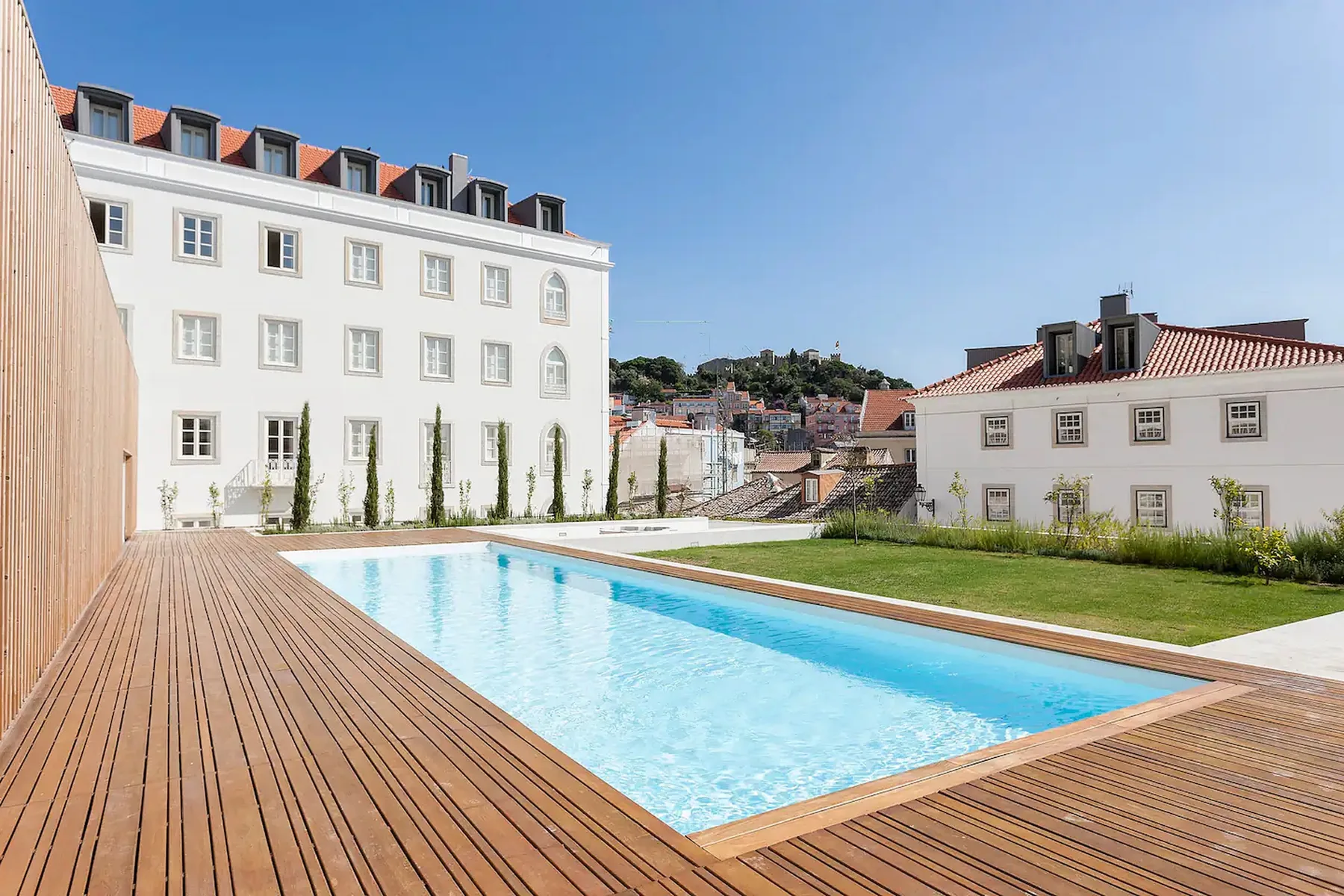 Una piscina rectangular al aire libre está junto a una tarima de madera y edificios blancos.