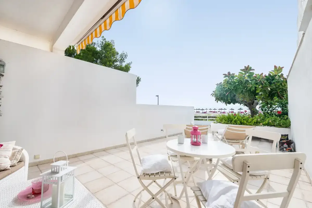 Un patio exterior con suelo de baldosas, asientos blancos, una mesa redonda y plantas.
