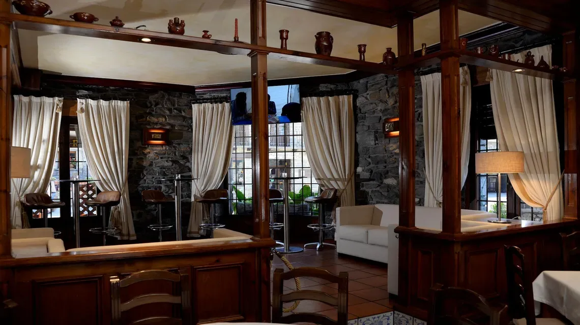 Interior del restaurante del hotel con molduras de madera oscura, paredes de piedra, cortinas y mesas.