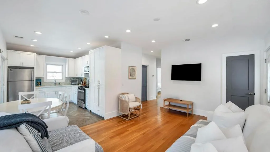 A bright open-plan living room and kitchen with white seating and wood flooring.