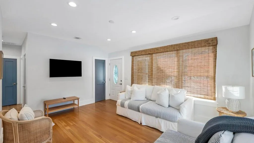 Una sala de estar con asientos blancos, un televisor en la pared y una ventana amplia.