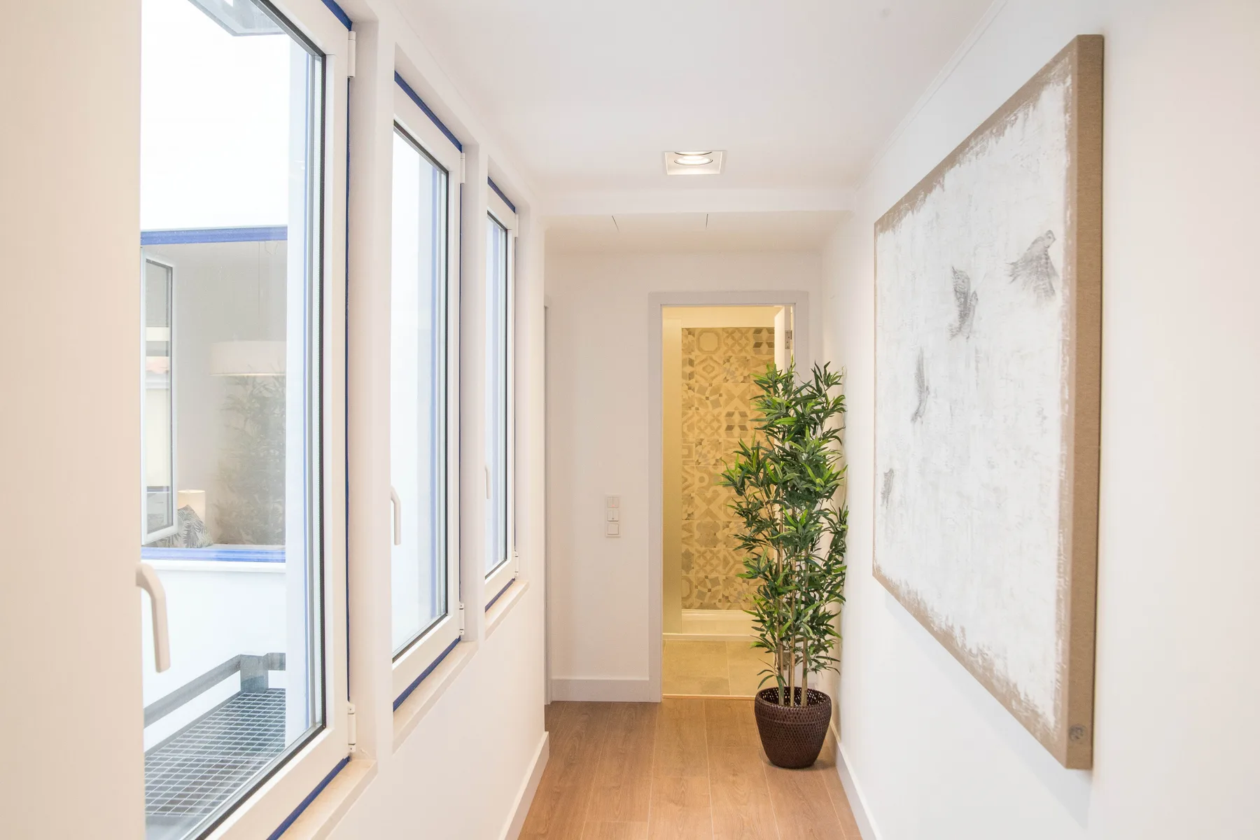 Un pasillo luminoso con ventanas grandes, una planta en maceta y arte en la pared.
