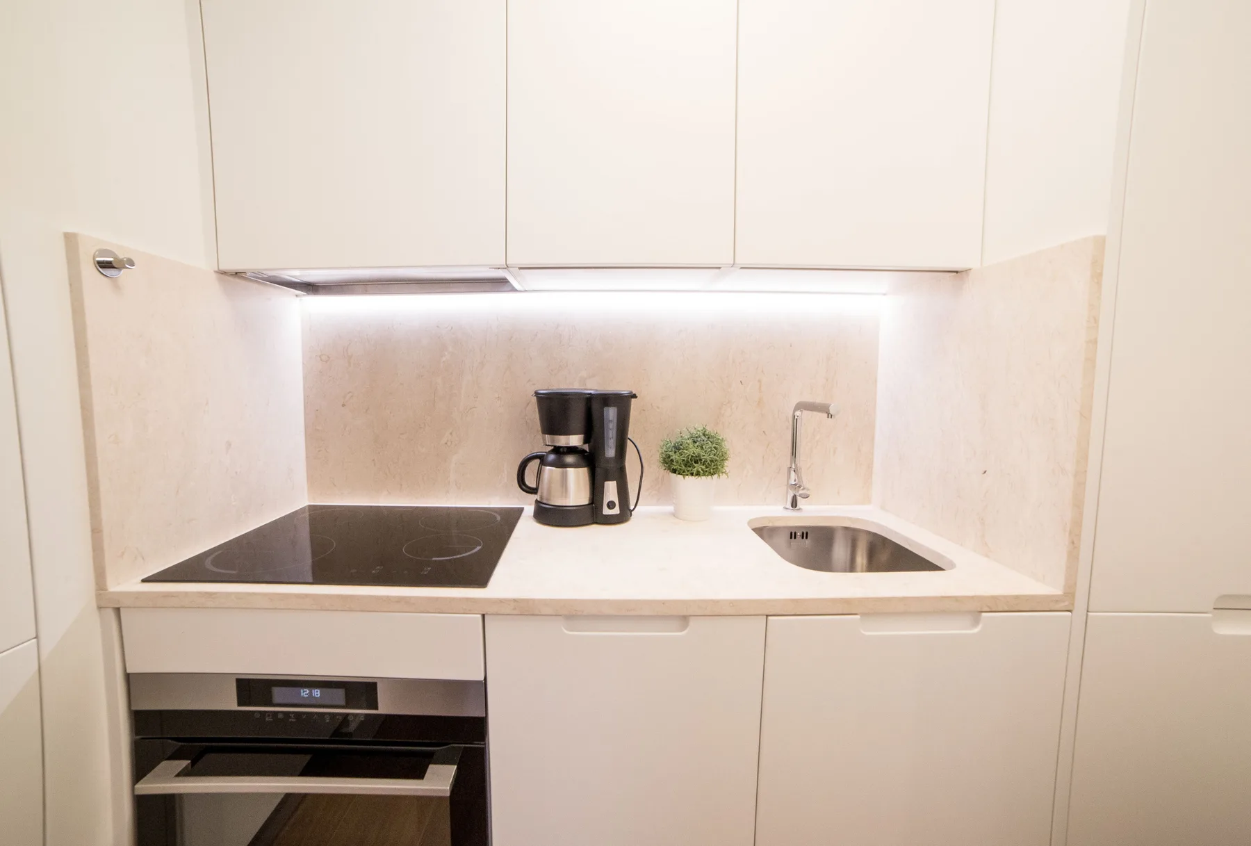 Una cocina compacta con placa de cocción, cafetera y fregadero bajo armarios blancos.