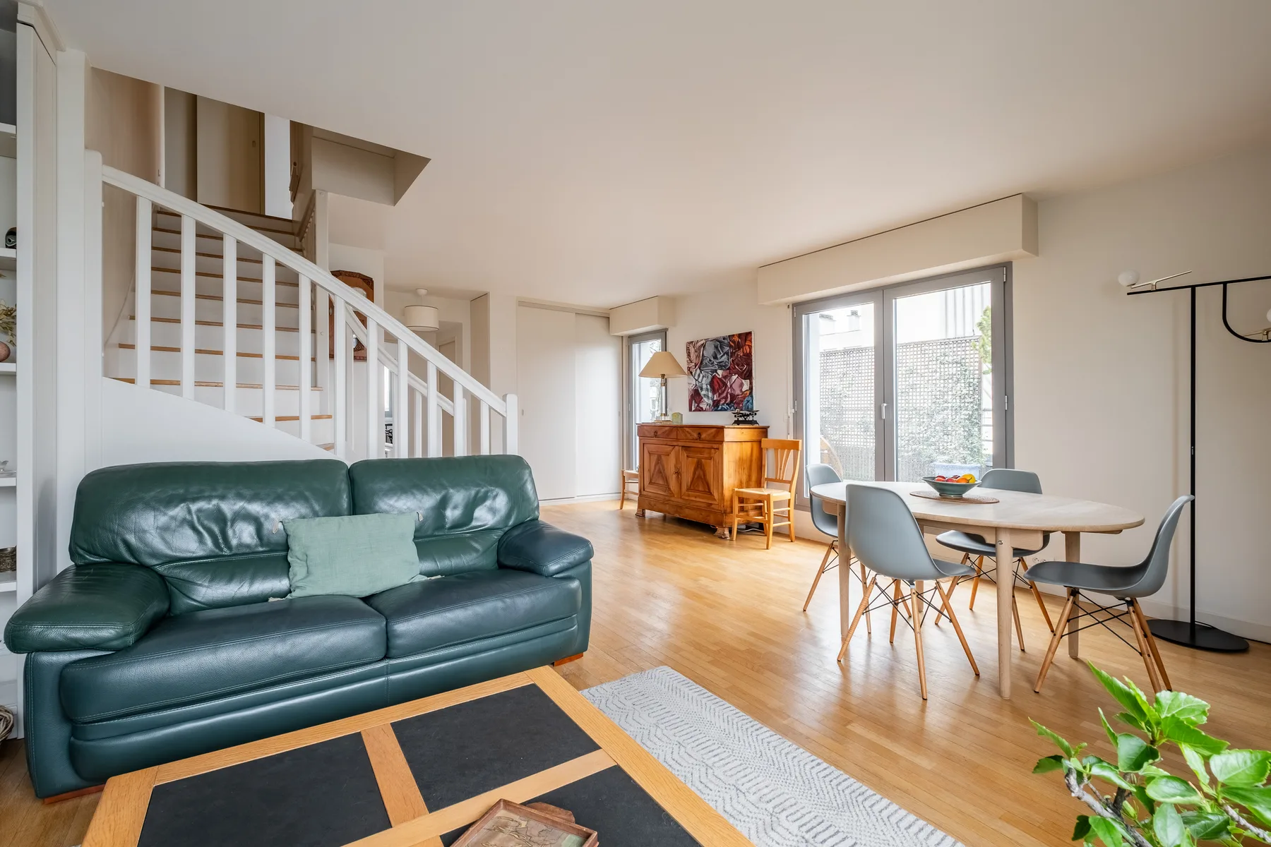 A bright living and dining area has green seating, a wooden table, and large windows.