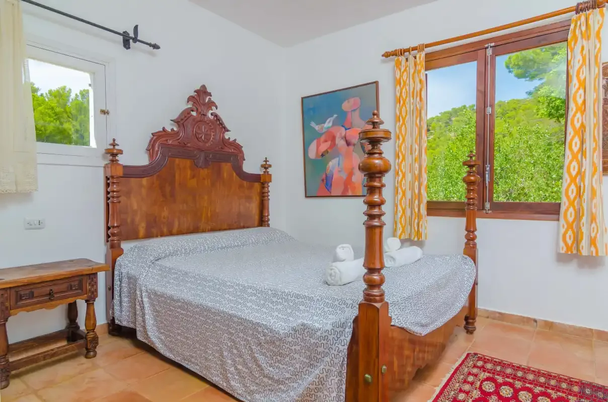 Un dormitorio con una cama de madera ornamentada, ropa de cama estampada y una ventana con vistas a la vegetación.