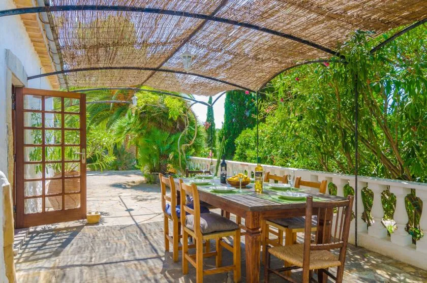 A shaded outdoor dining area with a wooden table and chairs beside lush greenery.