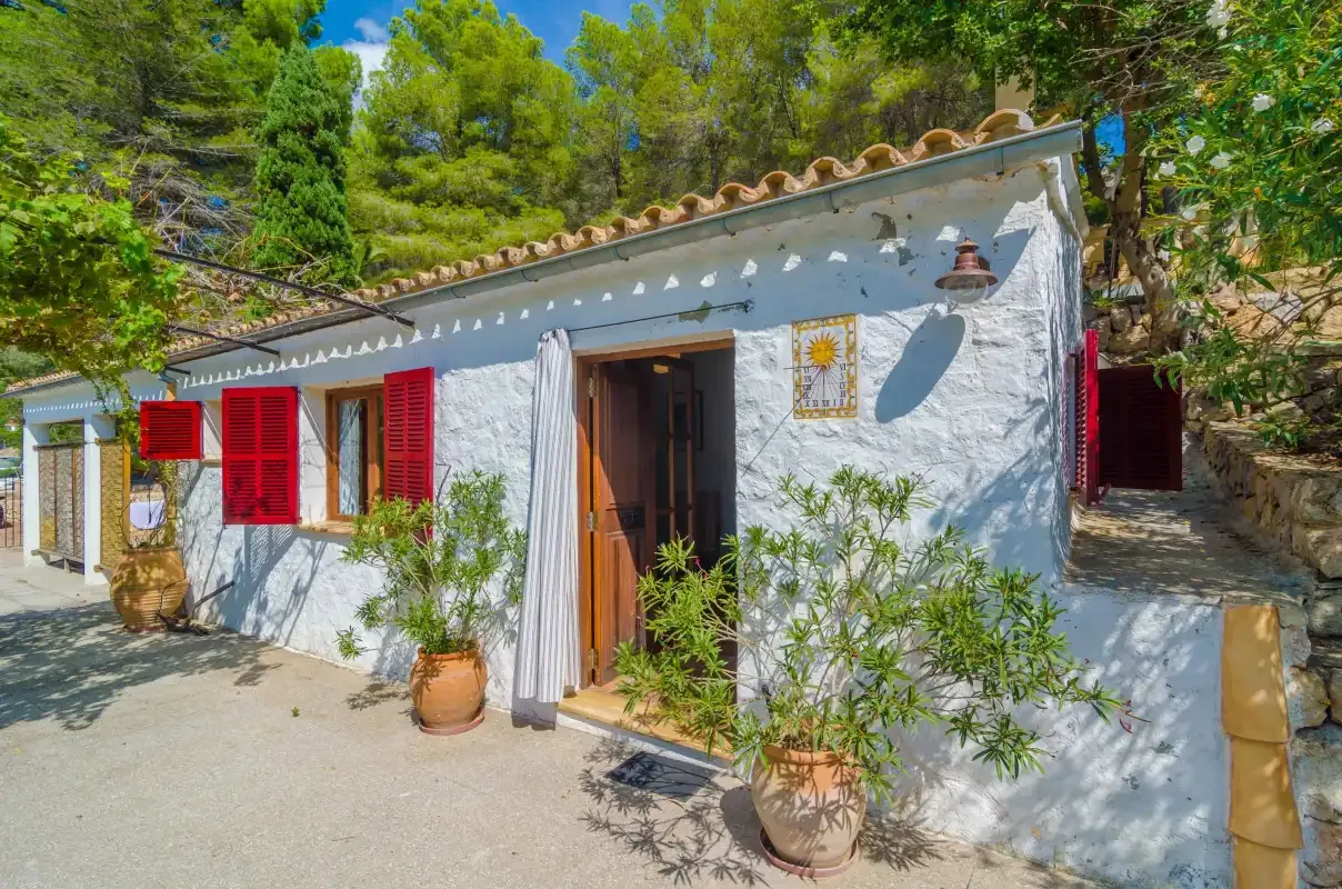 Una casa blanca con contraventanas rojas y plantas en macetas entre árboles.