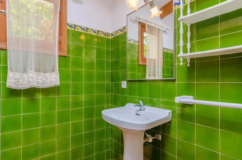 Un baño con azulejos verdes, lavabo con pedestal, espejo y ventana con cortina.