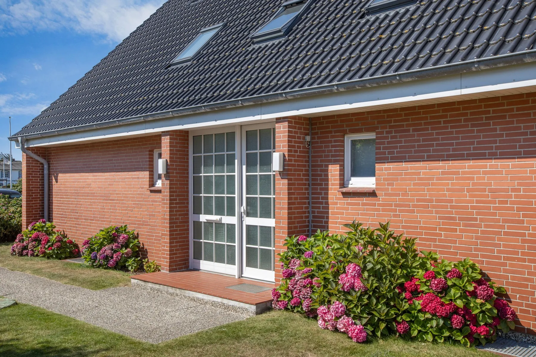 A brick house entrance with glass doors and flowering shrubs along the wall.