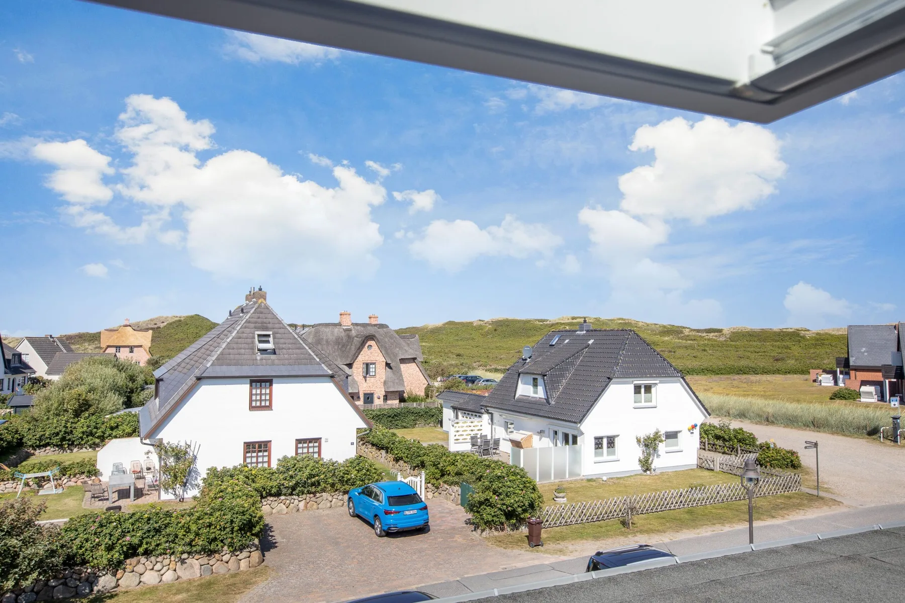 A sunny residential street with houses, parked cars, and grassy hills in the background.