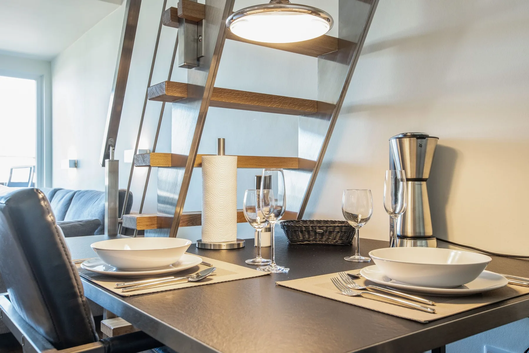 A dining table is set with plates and glasses beside an open staircase.
