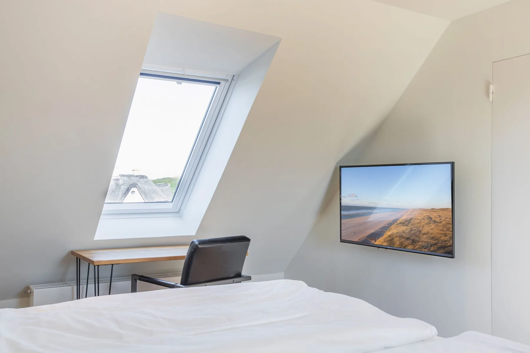 A bedroom with a bed, a desk beneath a skylight, and a wall-mounted television.