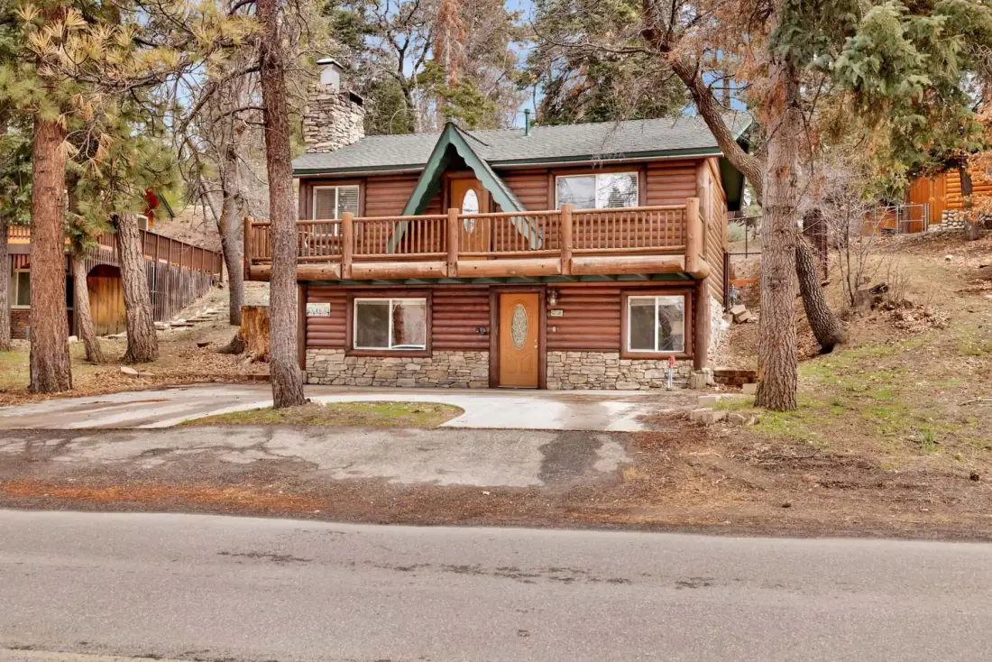 A two-story log cabin stands among trees beside a paved road.