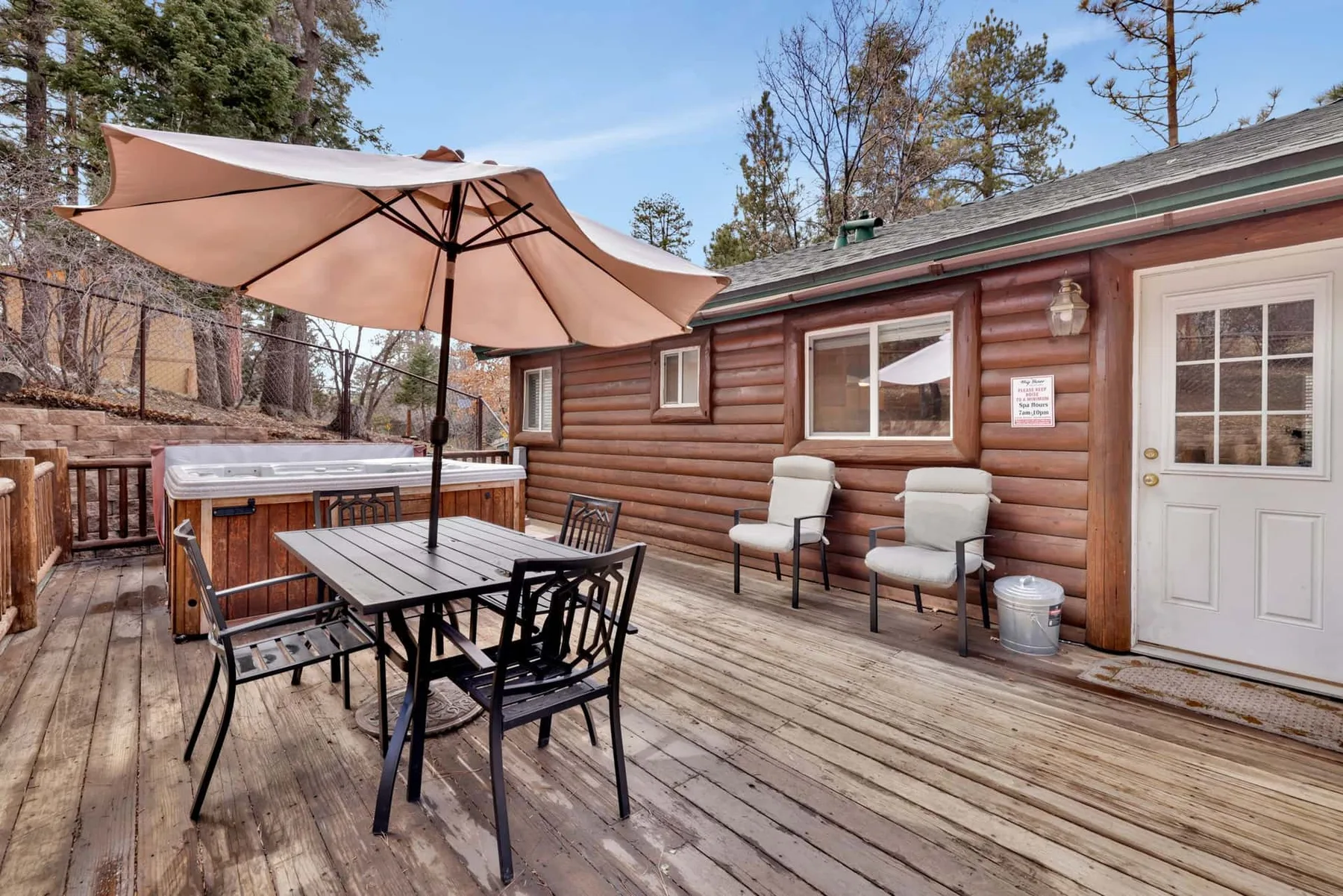 A wooden deck has an outdoor dining table, umbrella, and hot tub beside a cabin.