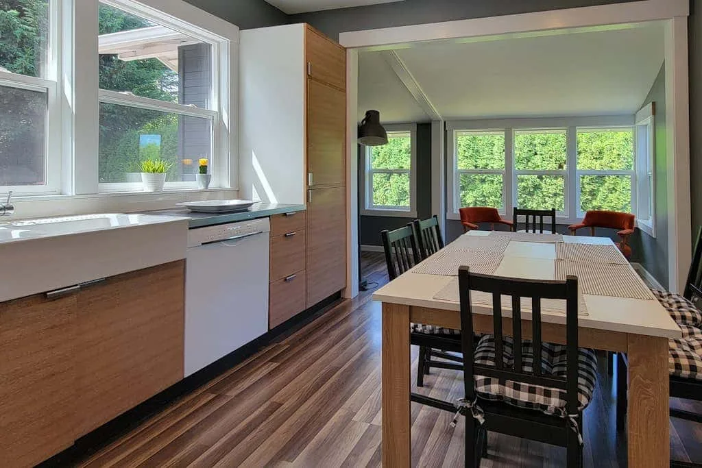 A dining area with a long table sits beside a kitchen counter and large windows.