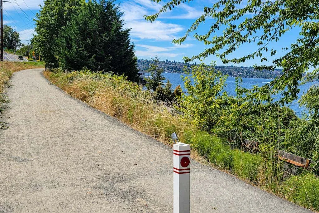 A paved path runs beside leafy vegetation, with a body of water visible beyond.