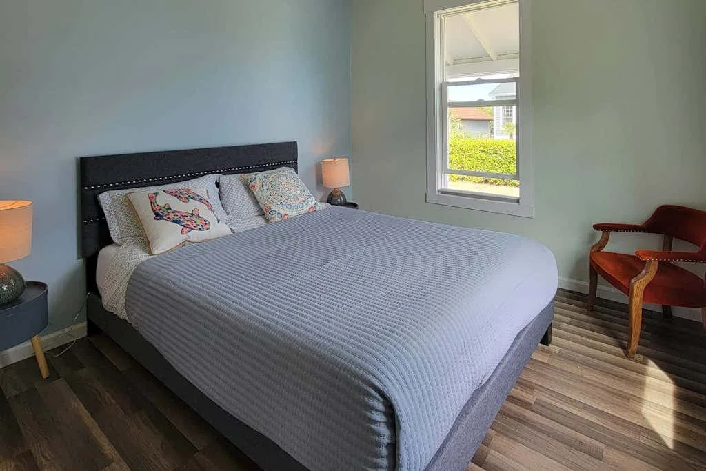 A bedroom with a large bed, patterned pillows, a window, and a wooden chair.