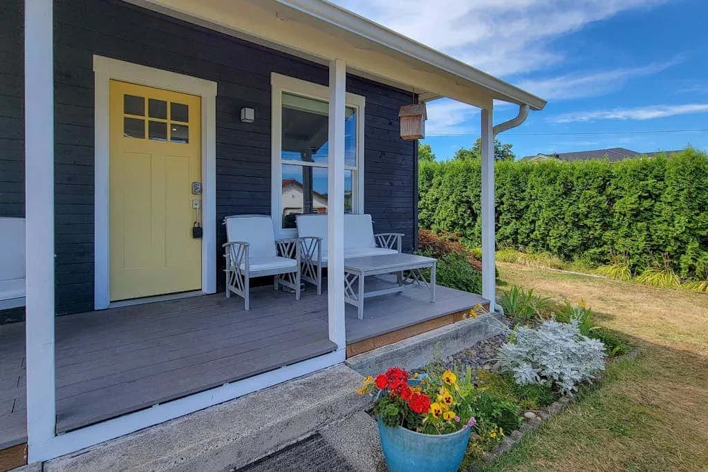 A covered porch with white seating faces a garden and hedge.