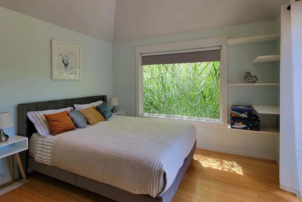 A bedroom with a neatly made bed beside a large window overlooking greenery.