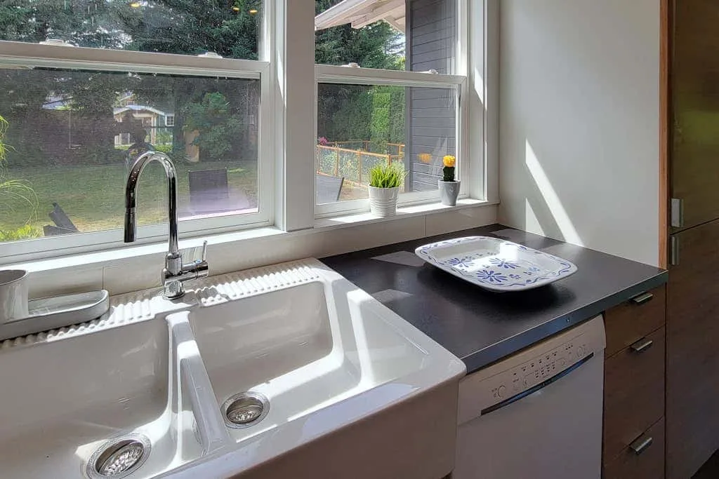 A kitchen sink and dark countertop sit beneath windows overlooking a garden.