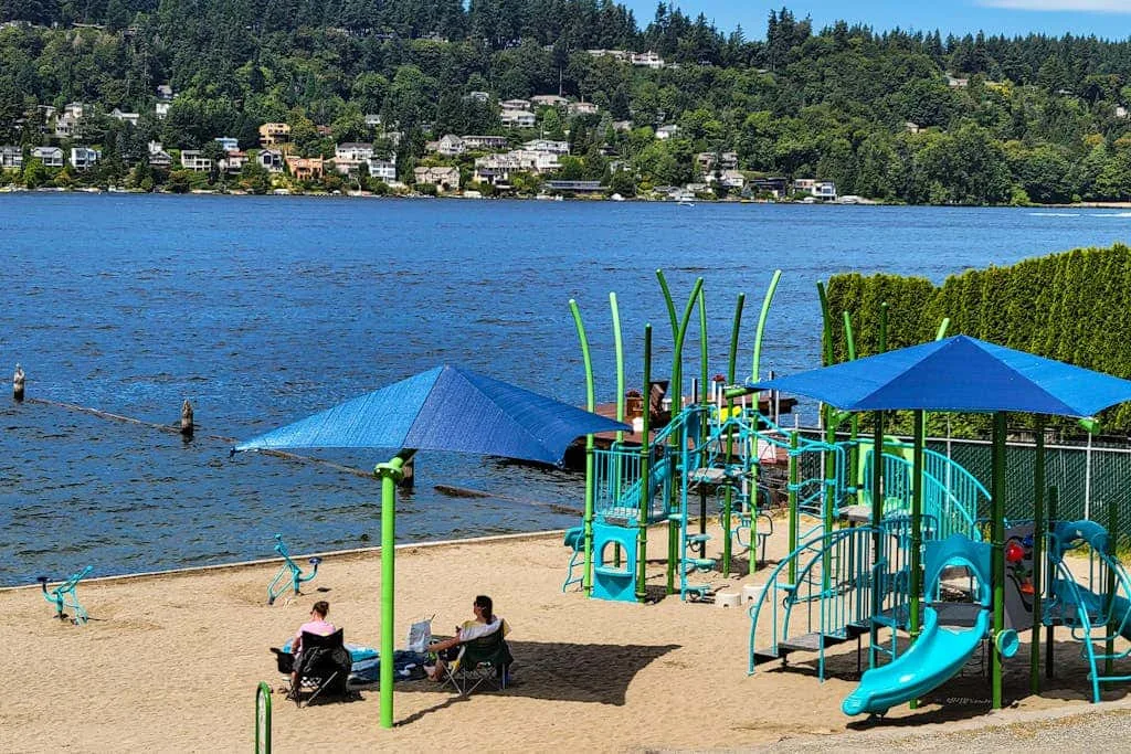A sandy beach playground sits beside a blue lake, with forested hills across the water.