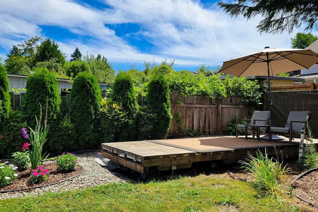 A landscaped backyard features a raised wooden deck, outdoor seating, and an umbrella.