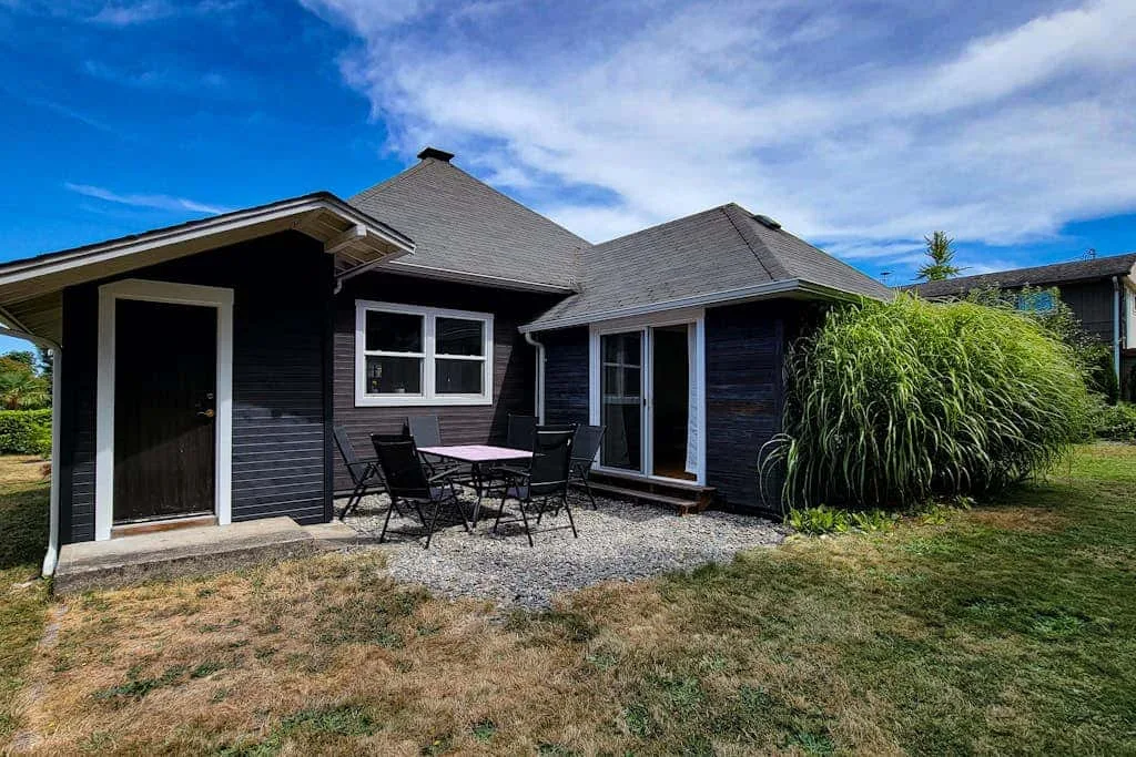 A dark-sided house has a small patio with outdoor seating beside the lawn.